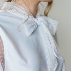 Puede incluir: Una blusa azul claro con un lazo blanco de lunares. La blusa tiene mangas de tela transparente con lunares.