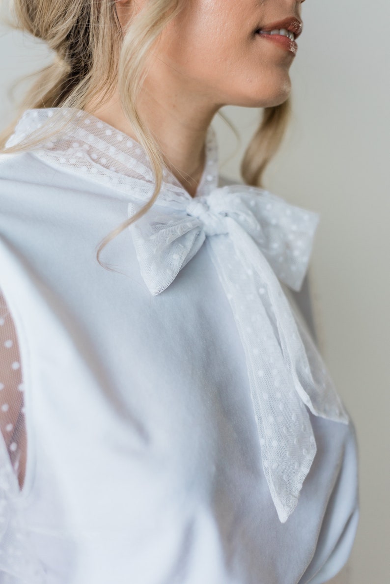 Puede incluir: Una blusa azul claro con un lazo blanco de lunares. La blusa tiene mangas de tela transparente con lunares.