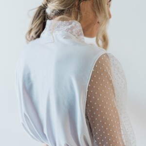 Puede incluir: Una blusa blanca con cuello alto y mangas largas. Las mangas est&aacute;n hechas de una tela transparente con lunares blancos.