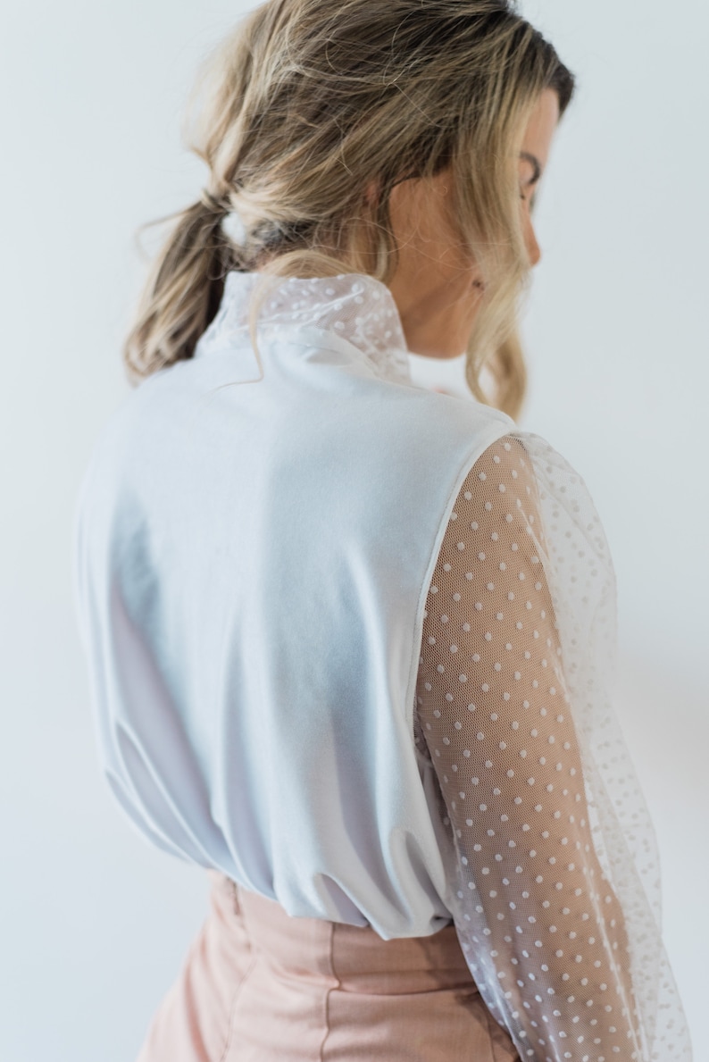 Puede incluir: Una blusa blanca con cuello alto y mangas largas. Las mangas est&aacute;n hechas de una tela transparente con lunares blancos.