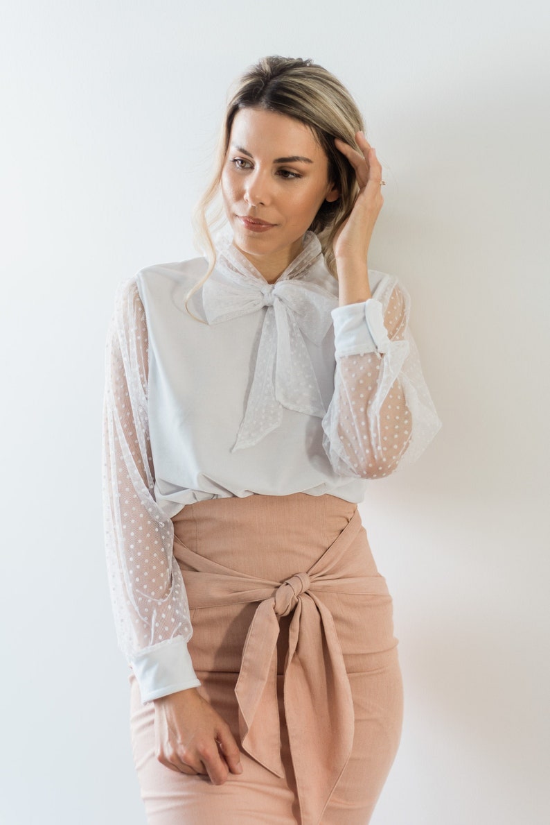 Puede incluir: Una mujer con una blusa blanca con una superposici&oacute;n de lunares transparentes y una corbata de lazo. Tambi&eacute;n lleva una falda rosa claro con una corbata de lazo.