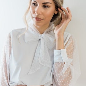 Puede incluir: Una blusa blanca con una superposici&oacute;n de tul de lunares y una corbata de lazo. La blusa tiene mangas largas con un pu&ntilde;o blanco y una falda rosa.