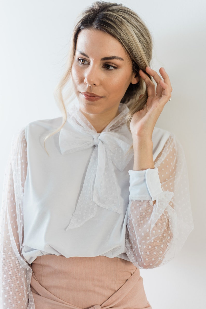 Puede incluir: Una blusa blanca con una superposici&oacute;n de tul de lunares y una corbata de lazo. La blusa tiene mangas largas con un pu&ntilde;o blanco y una falda rosa.