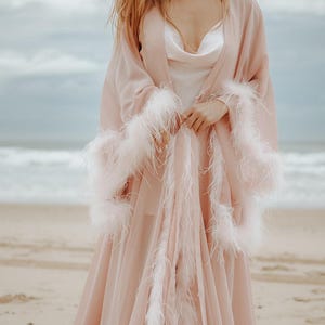 May include: A long, flowing, pale pink robe with white feather trim on the sleeves and edges. The robe is worn over a white slip dress. The image is taken on a beach with the ocean and sky in the background.
