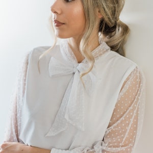 Puede incluir: Una blusa blanca con una superposici&oacute;n de lunares transparentes y un lazo en el cuello.