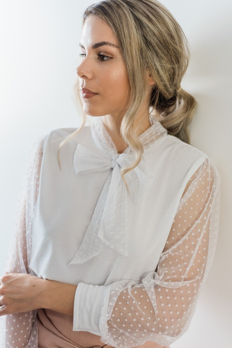 Puede incluir: Una blusa blanca con una superposici&oacute;n de lunares transparentes y un lazo en el cuello.