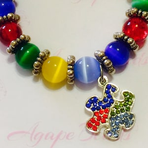 May include: A colorful beaded bracelet with a silver puzzle charm. The beads are red, green, yellow, blue, and light blue. The puzzle charm is decorated with red, green, blue, and light blue rhinestones.