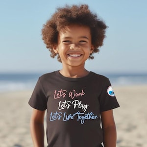 May include: A young person wearing a black t-shirt with the text "Let's Work Let's Play Let's Live Together" in pink, blue, and white lettering. The person is smiling and standing on a beach.