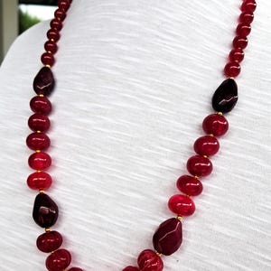 Può includere: Una collana lunga caratterizzata da una serie di perline rotonde e sfaccettate in varie tonalità di rosso intenso e bordeaux. Le perline sono infilate insieme a piccoli distanziatori color oro, creando un filo continuo di pietre rosse.