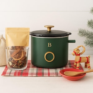 May include: A festive arrangement featuring a dark green slow cooker with gold accents, a glass of dried fruit and herbs, and a small straw goat figurine. A red and white plaid cloth and a small red bowl with a wooden spoon complete the scene.