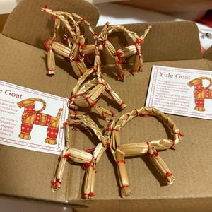 Peut inclure: Plusieurs chèvres de Yule en paille, décorations de Noël traditionnelles, sont présentées dans une boîte en carton. Chaque chèvre est faite de paille et attachée avec de la ficelle rouge. Deux cartes avec le texte "Yule Goat" sont également visibles.