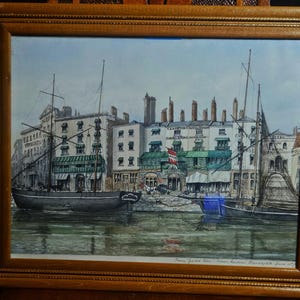 Puede incluir: Pintura de acuarela enmarcada de una escena portuaria con varios barcos atracados a lo largo de un muelle de piedra. Los edificios con toldos y letreros bordean la costa. La pintura está en un marco de madera.