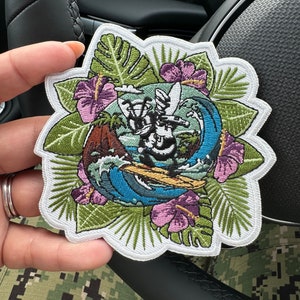 May include: Embroidered patch with a tropical theme. It features a blue and white wave, a palm tree, and a hibiscus flower. The patch is surrounded by green leaves.