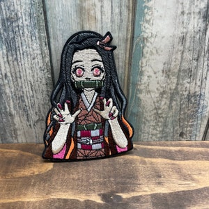 May include: Embroidered patch of a female anime character with long black hair, wearing a brown and red kimono with a green sash. She has pink eyes and is wearing a green and white mask. Her hands are raised with pink fingernails.