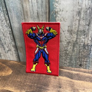 May include: Embroidered patch featuring a muscular superhero in a blue and red costume with yellow accents. The patch is rectangular with a red background and the character is flexing. The patch is likely for a jacket or bag.