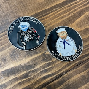 May include: Two black and silver round challenge coins with a white cartoon character on each side. The text "Let Fate Decide" is printed around the edge of each coin.