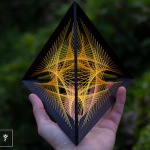 Könnte beinhalten: Eine schwarze pyramidenförmige String-Art-Skulptur mit einem geometrischen Muster aus gelbem und orangefarbenem Garn. Die Skulptur wird in der Hand einer Person gehalten.