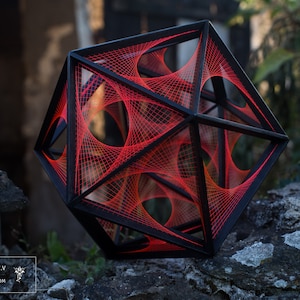 Peut inclure: Une sculpture géométrique en bois noir et en fil rouge. La sculpture a la forme d'un cube avec un motif complexe de fil tissé à travers lui. La sculpture est posée sur un rocher.