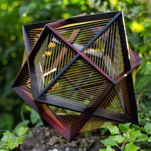 May include: A geometric wooden sculpture with a dark wood frame and gold string accents. The sculpture is in the shape of a cube with a complex pattern of intersecting lines.