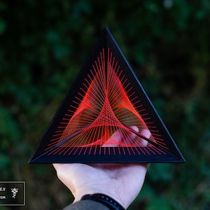 May include: A black triangular frame with a red string art design inside. The string art design is a geometric pattern of intersecting lines.