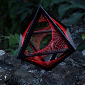 May include: A black geometric sculpture with a red wire mesh interior. The sculpture is made of wood and wire and is resting on a rock.