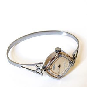May include: A silver-toned bangle watch featuring a square-shaped case with rounded corners. The watch face is cream-colored with black hands and blue decorative accents. The band is a rigid, open-style metal cuff with triangular cutouts.