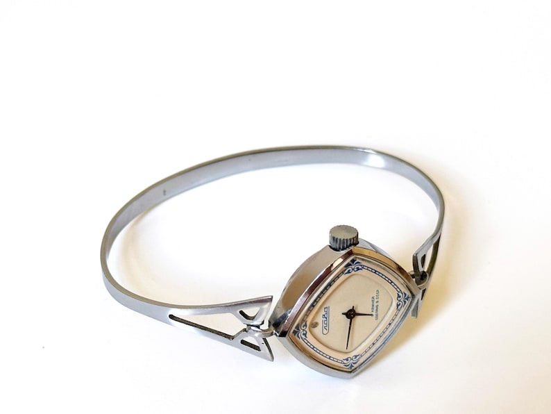 May include: A silver-toned bangle watch featuring a square-shaped case with rounded corners. The watch face is cream-colored with black hands and blue decorative accents. The band is a rigid, open-style metal cuff with triangular cutouts.