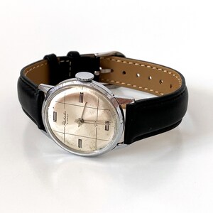 May include: A vintage wristwatch with a silver-colored case and a light-colored face featuring a grid pattern. The watch has black leather straps and a silver-colored crown. The watch face has the word "Raketa" printed on it.