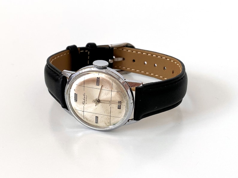 May include: A vintage wristwatch with a silver-colored case and a light-colored face featuring a grid pattern. The watch has black leather straps and a silver-colored crown. The watch face has the word "Raketa" printed on it.