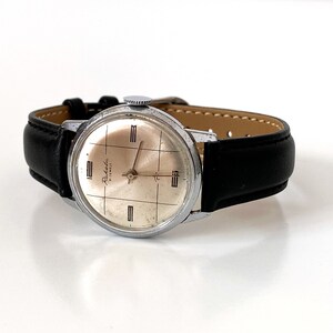 May include: A vintage Raketa watch with a silver-colored face and a black leather band. The watch face has a grid pattern and the words "Raketa" and "17 Jewels". The watch is a classic timepiece.