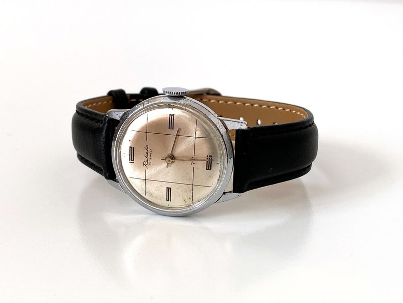 May include: A vintage Raketa watch with a silver-colored face and a black leather band. The watch face has a grid pattern and the words "Raketa" and "17 Jewels". The watch is a classic timepiece.