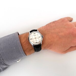 May include: A vintage wristwatch with a silver-toned case and a black leather band. The watch face is cream-colored with a grid pattern and silver hands. The watch is on a person's wrist, with a gray shirt sleeve visible.