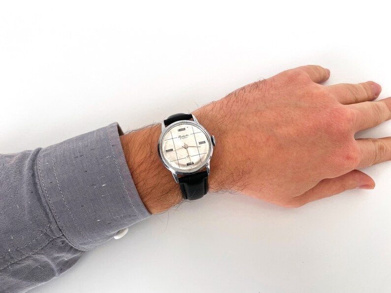 May include: A vintage wristwatch with a silver-toned case and a black leather band. The watch face is cream-colored with a grid pattern and silver hands. The watch is on a person's wrist, with a gray shirt sleeve visible.