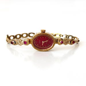 Op de afbeelding: Een vintage goudkleurig polshorloge met een rode ovale wijzerplaat en een bijpassende armband. Het horloge heeft kleine rode edelstenen en decoratieve bloemen- en geometrische patronen. Het horloge wordt op een witte achtergrond weergegeven.