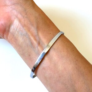 May include: A simple, polished silver-tone metal bangle bracelet with a smooth, flat surface and a thin, uniform width. The bracelet features a minimalist, open-ended design that encircles the wrist, showcasing a reflective, metallic finish.