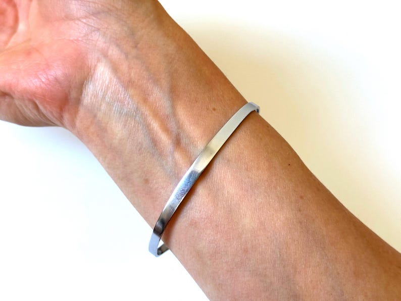 May include: A simple, polished silver-tone metal bangle bracelet with a smooth, flat surface and a thin, uniform width. The bracelet features a minimalist, open-ended design that encircles the wrist, showcasing a reflective, metallic finish.