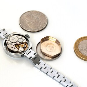 May include: A silver wristwatch with a round face and a silver metal strap. The watch is open, revealing the inner workings of the watch movement.
