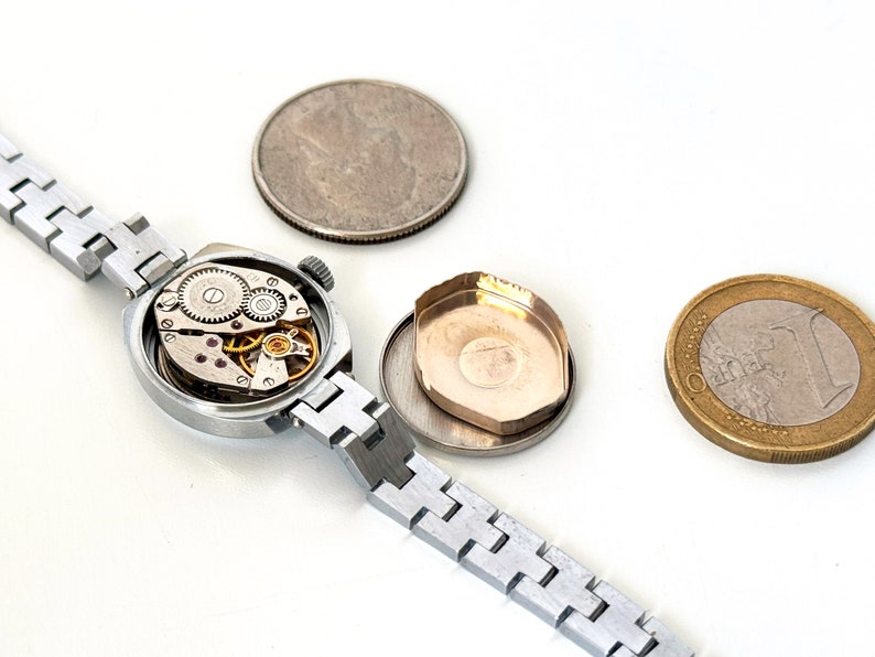 May include: A silver wristwatch with a round face and a silver metal strap. The watch is open, revealing the inner workings of the watch movement.