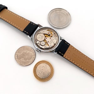 May include: An open-faced vintage wristwatch with a black leather band, revealing the intricate internal mechanisms. A quarter and a Euro coin are placed near the watch. The watch band is marked "GENUINE LEATHER".