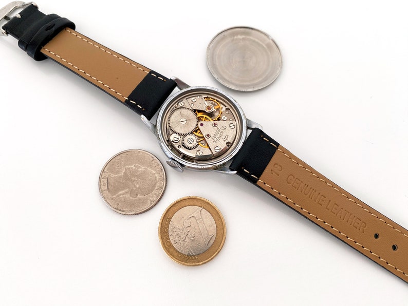 May include: An open-faced vintage wristwatch with a black leather band, revealing the intricate internal mechanisms. A quarter and a Euro coin are placed near the watch. The watch band is marked "GENUINE LEATHER".
