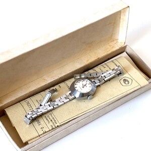 May include: A silver wristwatch with a white face and a silver metal strap. The watch is in a white box with a paper insert that has text in Russian.