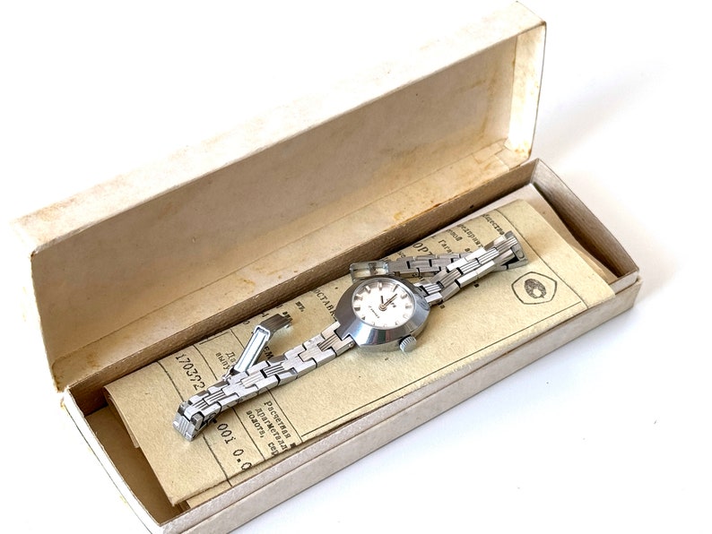 May include: A silver wristwatch with a white face and a silver metal strap. The watch is in a white box with a paper insert that has text in Russian.