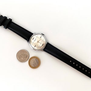 May include: A vintage wristwatch with a silver-colored face and black leather band. The watch face has a grid pattern. Two coins are next to the watch. The watch band is approximately 0.75 inches wide.