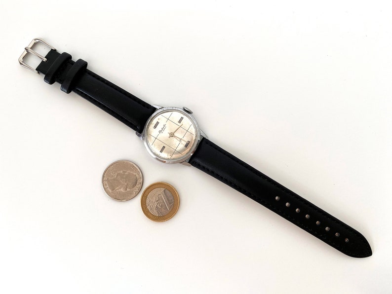 May include: A vintage wristwatch with a silver-colored face and black leather band. The watch face has a grid pattern. Two coins are next to the watch. The watch band is approximately 0.75 inches wide.