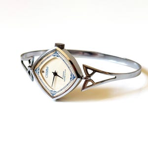 May include: A silver-toned bangle watch with a square-shaped face featuring a cream-colored dial, black hands, and blue decorative accents. The watch includes a crown at the top and a rigid, open-work metal band with geometric triangular cutouts.