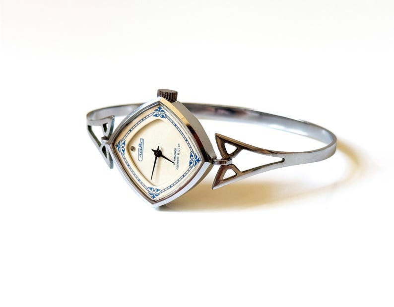 May include: A silver-toned bangle watch with a square-shaped face featuring a cream-colored dial, black hands, and blue decorative accents. The watch includes a crown at the top and a rigid, open-work metal band with geometric triangular cutouts.