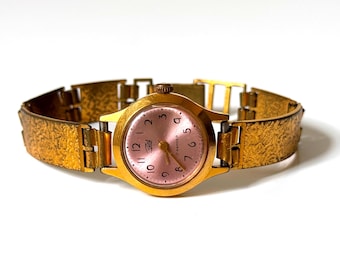 May include: A gold-tone wristwatch featuring a round case with a metallic pink dial, black Arabic numerals, and a textured link bracelet. The watch face displays the text Zaria and 17 kamney in Cyrillic script with thin gold hands.