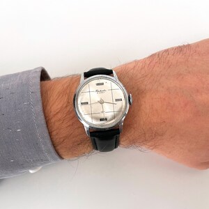 May include: A vintage silver-toned wristwatch with a black leather band. The watch face is silver with black grid lines and the word "Raketa" is printed on the face. The watch is on a person's wrist.