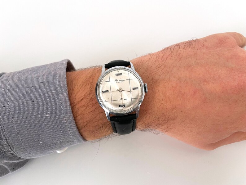 May include: A vintage silver-toned wristwatch with a black leather band. The watch face is silver with black grid lines and the word "Raketa" is printed on the face. The watch is on a person's wrist.