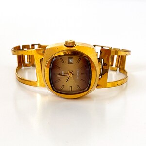 Puede incluir: Un reloj de pulsera dorado con una esfera cuadrada y una esfera marrón. El reloj tiene una pulsera de metal dorado con un diseño único.